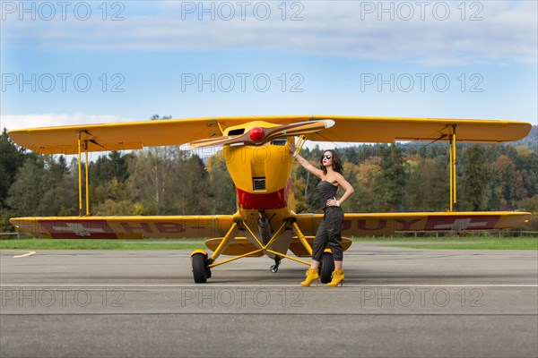 Young woman with sunglasses in overall and boots posing in front of double-decker airplane