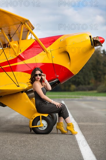 Young woman with sunglasses in overall and boots posing in front of double-decker airplane