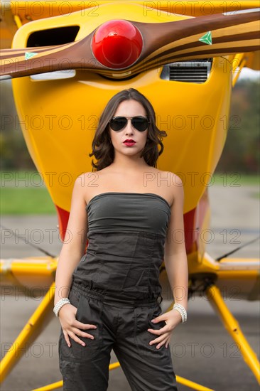 Young woman with sunglasses in overall and boots posing in front of double-decker airplane