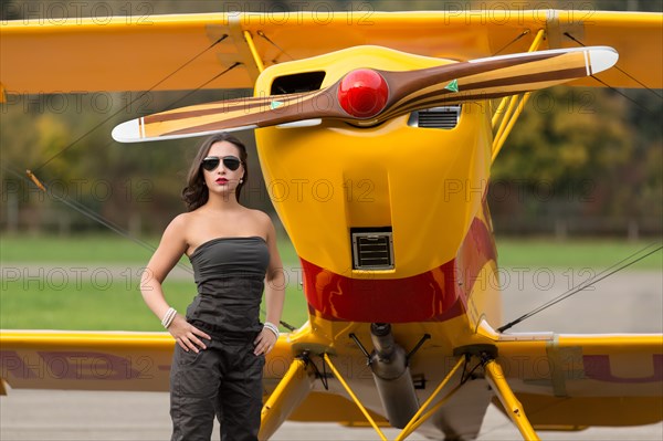 Young woman with sunglasses in overall and boots posing in front of double-decker airplane