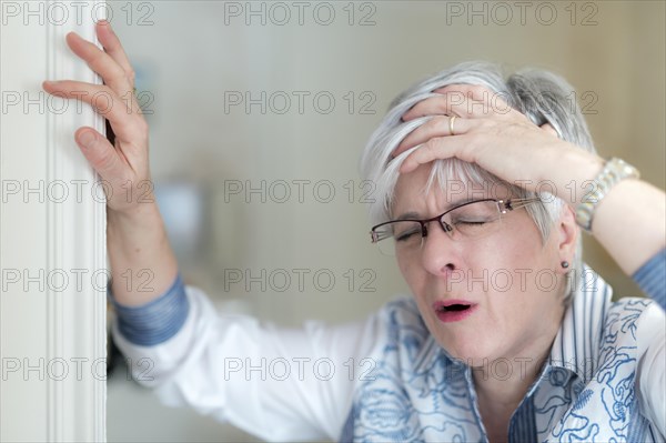 Senior woman with a headache holds her hand on her forehead and props herself on the door frame