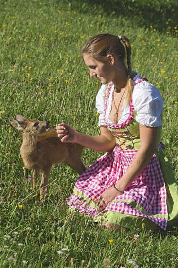 Woman in dirndl with a tame fawn in a flower meadow