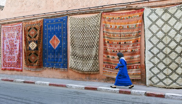 Moroccan woman walking past carpets hanging on wall