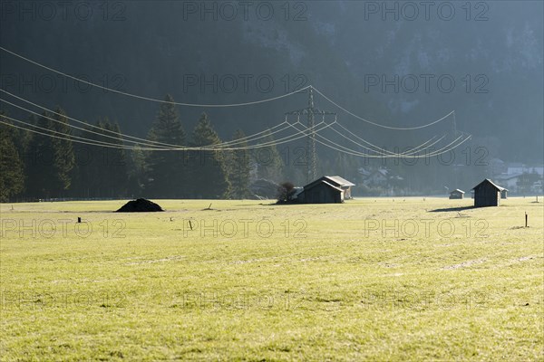 An electricity line is crossing a grean meadow with some wooden huts in the morning light
