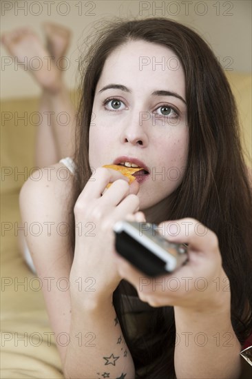 Young woman watching TV on the couch with chips and the remote control in hand