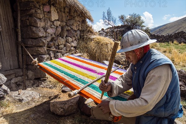 Old Indio man with helmet weaving a colorful rug with a simple loom