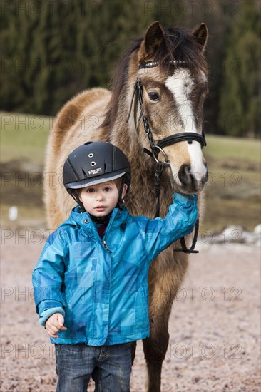 Young child wearing a riding helmet standing beside a pony