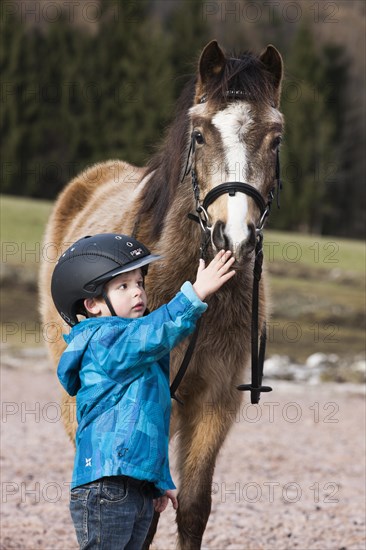 Young child wearing a riding helmet standing beside a pony