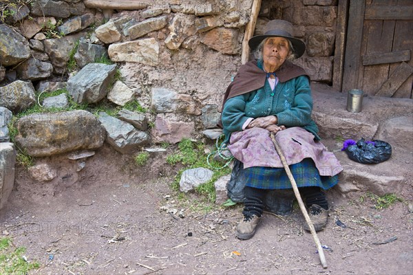 Mature Quechua Indian woman wearing a hat sitting on a stone in front of a doorway
