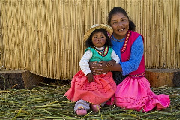 Friendly woman and a girl of the Uro Indians wearing traditional dress sitting in front of a reed hut