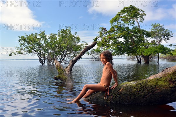 Girl sitting on a tree trunk of a giant rainforest tree on the banks of the Amazon and Rio Solimoes