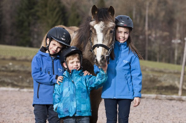 Three children wearing riding helmets standing beside a pony