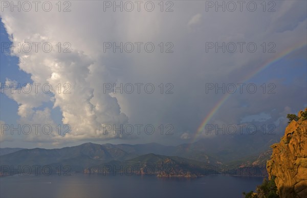 The typical bizarre red rocks of the Calanche of Piana and the mediterranean sea at the Gulf of Porto in the background below a rainbow and some clouds. The Calanche of Piana is in the western part of the island Corsica