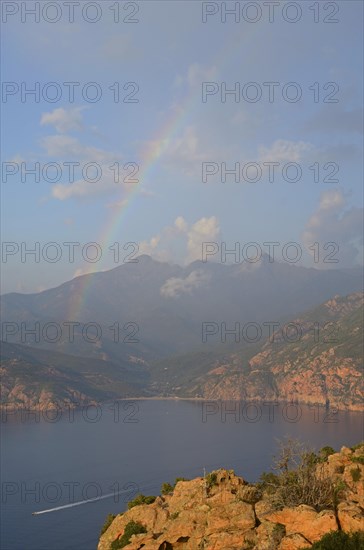 The typical bizarre red rocks of the Calanche of Piana and the mediterranean sea at the Gulf of Porto in the background below a rainbow and some clouds. The Calanche of Piana is in the western part of the island Corsica