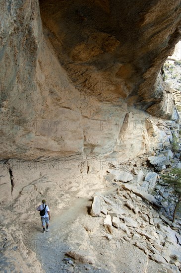 Local hiker walking under a rock overhang on the path to the abandoned settlement of Sap Bani Khamis on the western flank of the Grand Canyon of Oman in the Wadi Nakhar at the foot of the Jebel Shams summit