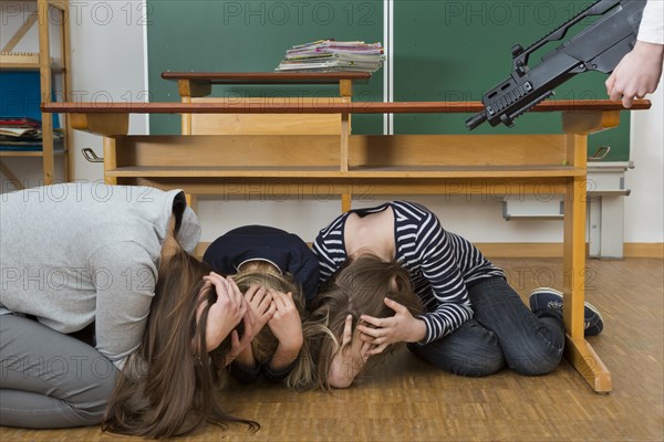 Teacher and school children crouching under a desk in front of a gunman