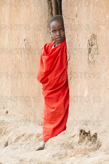 Maasai child wearing traditional dress looking out of a mud hut