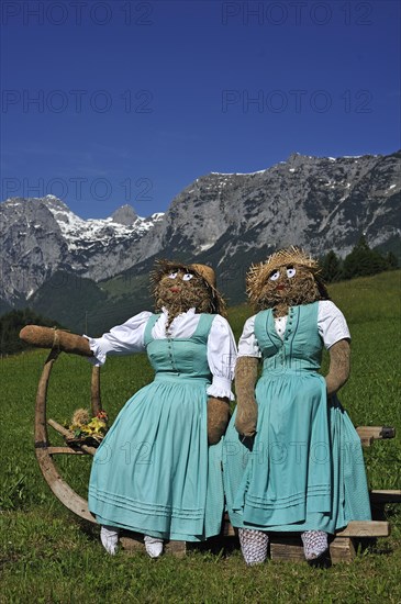 Two straw dolls or figures in dirndl dresses sitting on a sledge with long horn-shaped runners on a meadow