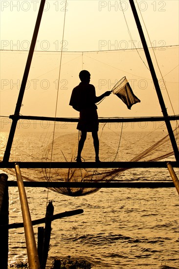 Fisherman holding a landing net in front of a Chinese fishing net at sunrise