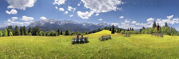 Hikers relaxing on a bench in the middle of a flowery meadow with panoramic views towards the Wetterstein Massif with the Alps and Zugspitze Mountain