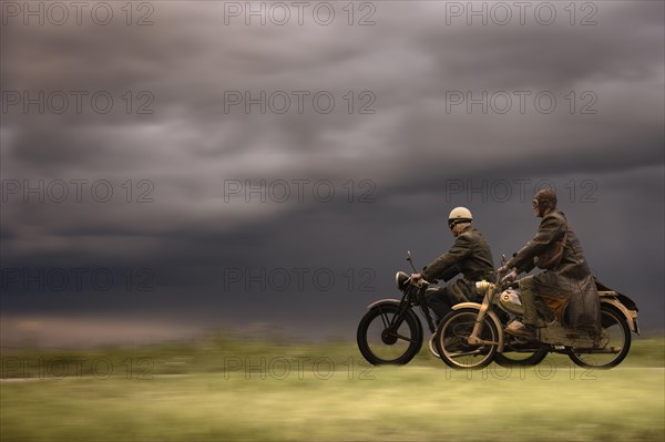 Two vintage motorcyclists in front of a dramatic cloudy sky