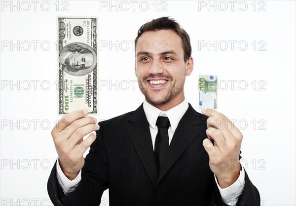 Smiling young man wearing a suit holding a large 100 dollar bill and a small 100 euro banknote