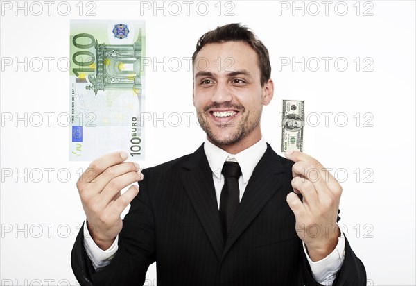 Smiling young man wearing a suit holding a large 100 euro banknote and a small 100 dollar bill