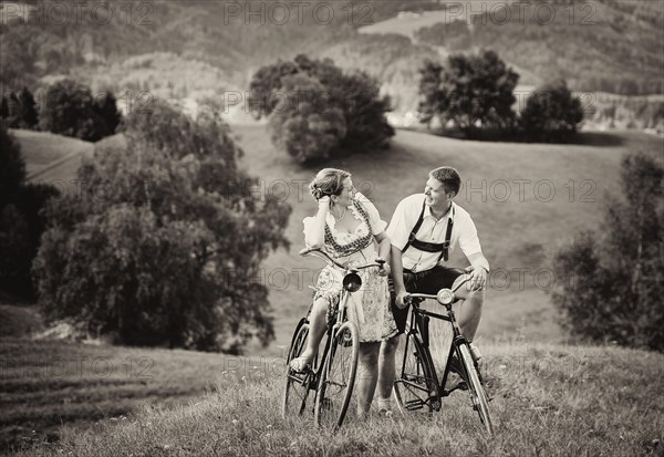 Man wearing leather pants and a woman wearing a dirndl on old bicycles within a natural landscape
