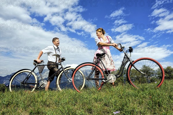 Man wearing leather pants and a woman wearing a dirndl with old bicycles within a natural landscape