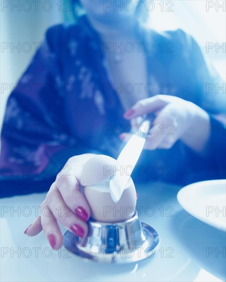 Woman wearing a bath robe slicing off the top of a breakfast egg with a knife