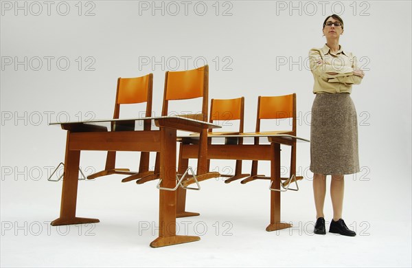 Strict female teacher standing next to old school desks with orange chairs