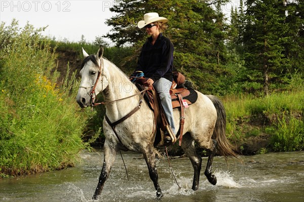 Cowgirl riding a gray horse through a river with splashing water