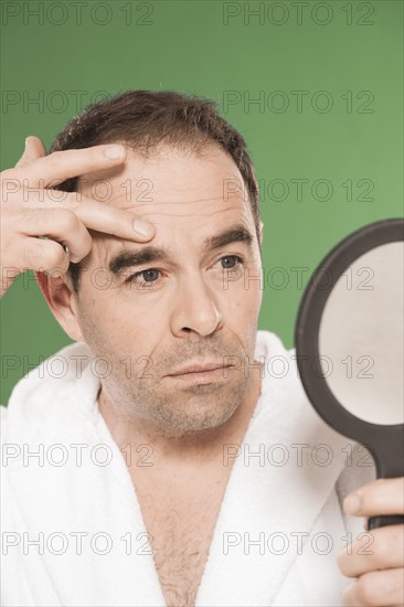 Man wearing a bathrobe observing himself critically in a hand mirror