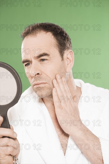 Man wearing a bathrobe observing himself critically in a hand mirror