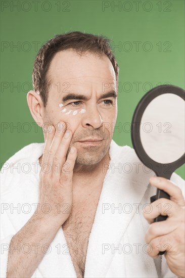 Man with eye cream observing himself critically in a hand mirror