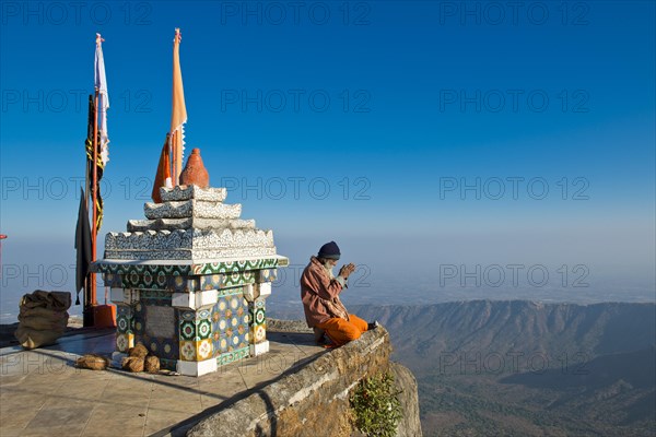Sadhu or holy man sitting with his hands folded in prayer on the precipice in front of a Hindu temple shrine with prayer flags