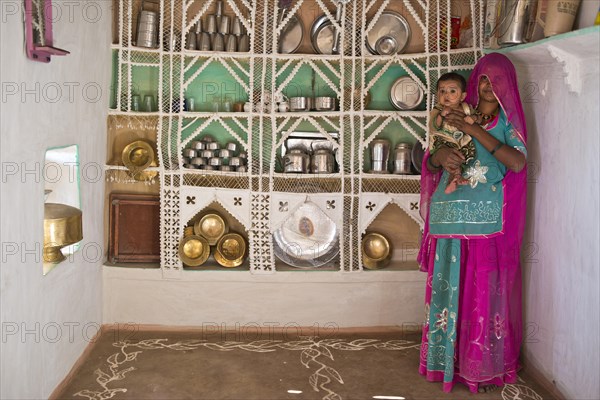 Young woman in colourful sari proudly standing with a child on her arm in front of a traditional kitchen shelf built from wire and sticks