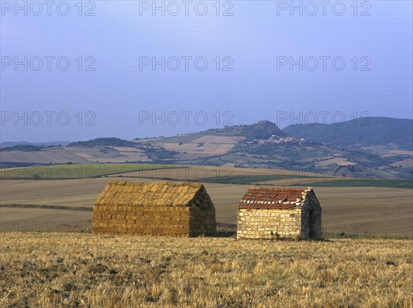 Bales of straw stacked in the shape of a house next to a little stone house