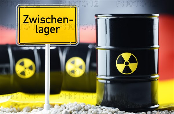 Three barrels of nuclear waste in front of a German flag and a town sign with the name "Zwischenlager"