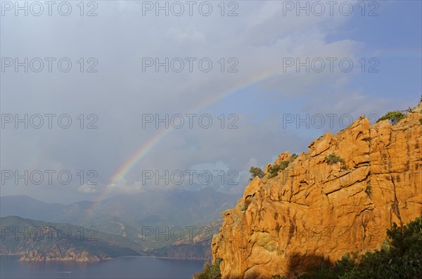 The typical bizarre red rocks of the Calanche of Piana and the mediterranean sea at the Gulf of Porto in the background below a rainbow and some clouds. The Calanche of Piana is in the western part of the island Corsica