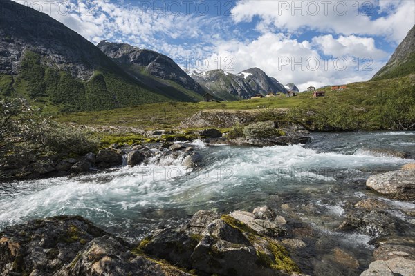 Torrent foaming between boulders in front of an alm or alpine meadow with huts and mountains