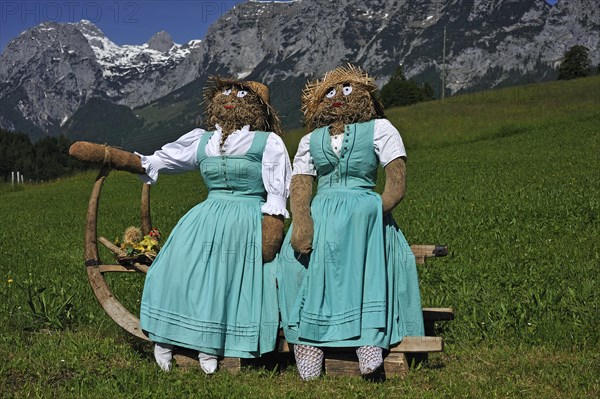Two straw dolls or figures in dirndl dresses sitting on a sledge with long horn-shaped runners on a meadow