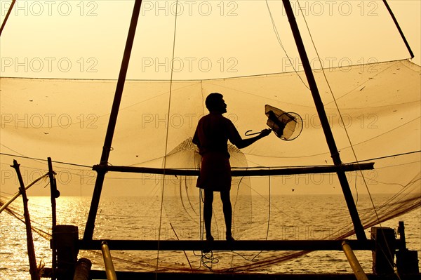 Fisherman holding a landing net in front of a Chinese fishing net at sunrise