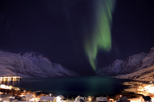 Aurora over a fjord with snow-covered mountains and an illuminated village