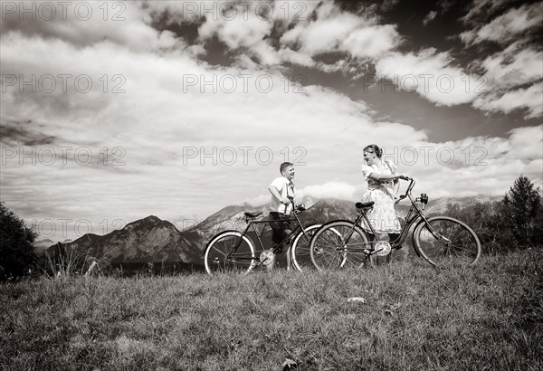 Man wearing leather pants and a woman wearing a dirndl with old bicycles within a natural landscape