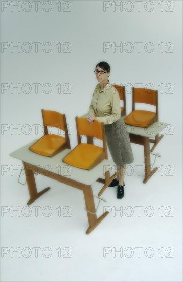 Strict female teacher standing at old school desks with orange chairs