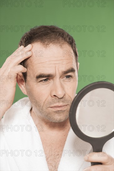 Man wearing a bathrobe observing himself critically in a hand mirror