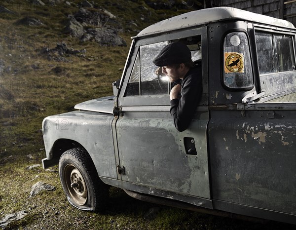 Man with a hat sitting in the driver's seat of an old car noticing a flat front left tyre