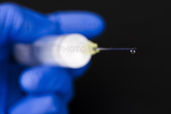 A hand with a blue medical glove is holding a syringe with a hypodermic needle and a drop of medicine