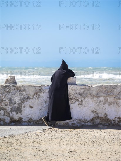 A Berber with black djellaba leaning against the harbor wall